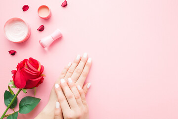 Young, perfect woman hands with nail polish bottle, cream jars and red rose on pink table. Pastel color. Care about nails and clean, soft, smooth skin. Empty place for text. Copy space. Top down view.