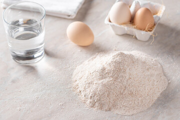 baking background with ingredients for kneading dough. a pile of whole grain flour, eggs, a glass of water on the kitchen table. place for text. horizontal image. selective focus