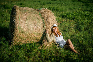 Naklejka na ściany i meble Beautiful girl is resting after work. girl on the field with hay. Woman near a sheaf of hay in a field. Rural life. Holidays in the village