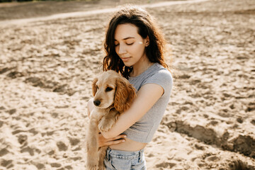Young brunette woman standing on sand on a beach, holding her little dog, cocker spaniel breed puppy.