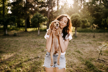 Young brunette woman holding her little dog, cocker spaniel breed puppy, outdoors, in a park.