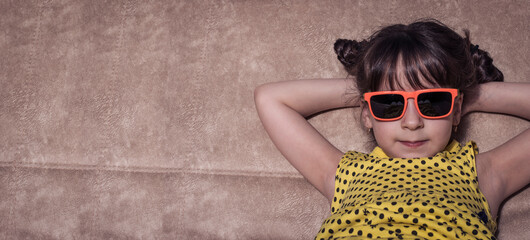 A cheerful little girl in orange sunglasses and a yellow dress with glasses, with her hands behind...