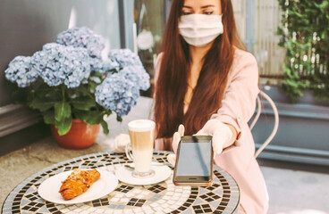 Portrait beautiful stylish woman in medical face mask use smart phone for contactless payment in outdoor french cafe. Young girl in protective face mask drink coffee, pay with NFC technology. COVID-19