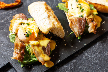 Sandwich with Eggs Benedict, bacon and salad on black stone background.