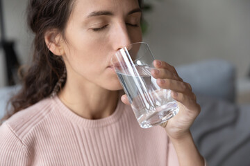 Close up beautiful young woman with closed eyes drinking pure mineral water, sitting on couch, attractive girl holding glass, healthy lifestyle and good daily habit concept, natural beauty