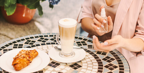Closeup stylish girl use antibacterial antiseptic gel on hand, drink coffee in french outdoor cafe. Beautiful woman apply hand sanitizer on restaurant terrace. Personal protection, hygiene, safety