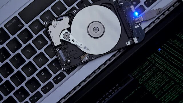 Hard drive lies on the laptop keyboard, flashes and disk spins. Work of hacked hard drive inside. Hacker breaks sensitive data from a hard drive. Against the background, lines of code and data flash. 