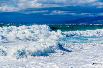 Storm waves in white foam rush in rows along the Tsemesskaya Bay. Blue sky and green sea. Dangerous and dramatic. In the background, mountains, multi-storey buildings of the city and port.