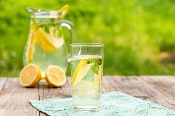 A glass of cold lemonade with lemon and mint on the background of a jug and green greenery with space to copy.