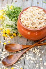 Hot oatmeal porridge in a clay pot with a bouquet of wild flowers and spoons on a wooden surface. Village Breakfast.
