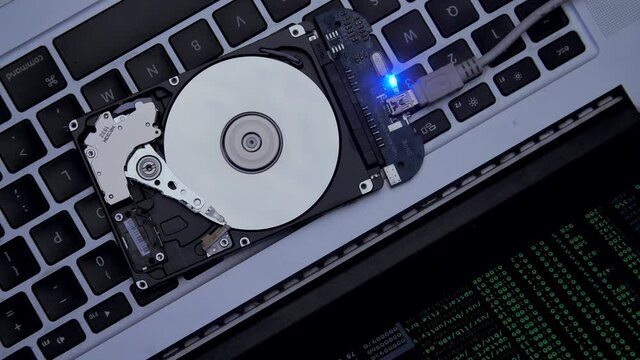 Hard drive lies on the laptop keyboard, flashes and disk spins. Work of hacked hard drive inside. Cybersecurity. Government secret data hacking technology. Lines of code and data flash on background