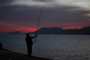 fishing at sunset with red horizon and a calm sea. Low light image showing the silhouette of the fisherman as well as the mountains