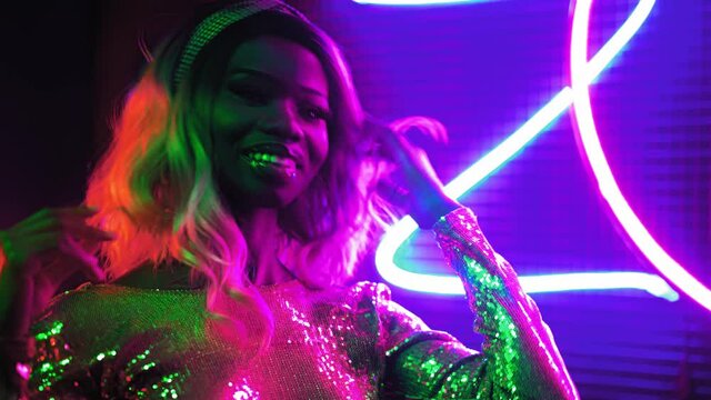 A ender African girl dance in 80s style posing on the camera in neon lighting in a silver sequin dress on a background of a round silver colored background. Prores 422.