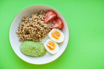 Bowl of oatmeal porridge with avocado, tomato, boiled egg and sesame on green background. Oatmeal with vegetables. Table top view in flat lay style. Hot and healthy breakfast and diet food