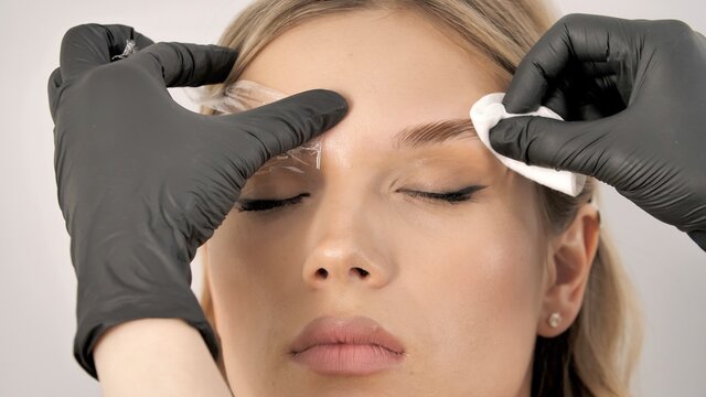 Stylist's hands in black gloves wipe the face of the model with a cotton pad. Beautiful attractive female face of a blonde well-groomed woman or lady. Styling and lamination of eyebrows. 