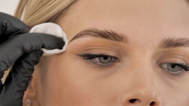 Stylist's hands in black gloves wipe the face of the model with a cotton pad. Beautiful attractive female face of a blonde well-groomed woman or lady. Styling and lamination of eyebrows. 
