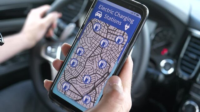 Closeup on a driver's hand holding a smartphone mobile device from inside his vehicle,using an app to search EV charging stations  to charge his electric car