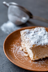 Cremșnit sitting on top of a plate. Is made out of filo pastry of usually three layers with cream on top and crumble sprinkled on it.