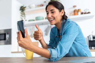 Beautiful young woman making video call and showing thumb up to smartphone during morning coffee at home.