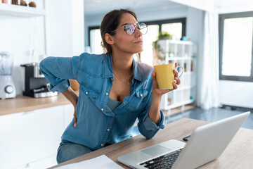 Tired business woman with back pain looking uncomfortable while working from home on laptop.