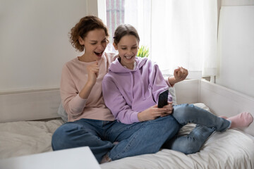 Happy Caucasian mom and teen daughter sit relax on bed at home have fun using cellphone together, overjoyed mother and teenage girl child play application game on modern smartphone gadget