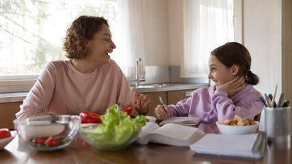 Smiling teen schoolgirl study prepare homework write in notebook at kitchen, young mom cooking chop...