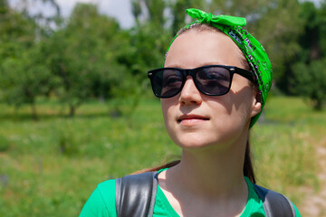 Portrait of a pretty hipster girl in sunglasses close up, green shirt, bandana standing on nature background on sunny day. Positive emotional caucasian young long haired lady smiles and looks on a sky