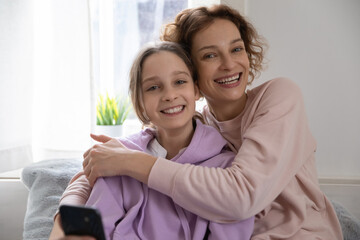 Portrait of happy young Caucasian mother and teen daughter sit relax at home hugging and cuddling, smiling mom and teenage girl child embrace, show love and care, family bonding concept