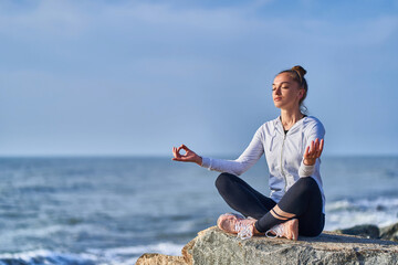 Fototapeta na wymiar Serene tranquil one woman in lotus position meditates and relaxes on a cliff against the background of the sea and clear blue sky. Calm concept and balance of mental health