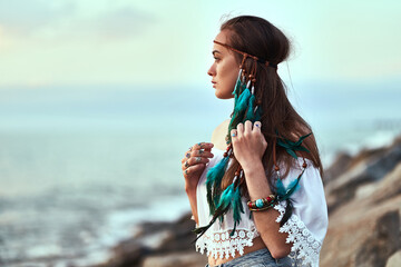 Portrait of attractive hippie woman wearing blue feathers in long hair, jewelry and white blouse at seashore. Boho chic style © Goffkein