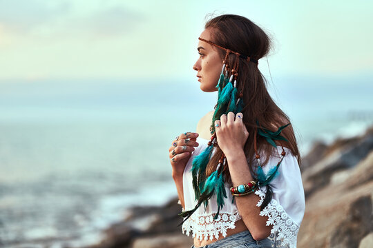 Portrait of attractive hippie woman wearing blue feathers in long hair, jewelry and white blouse at seashore. Boho chic style