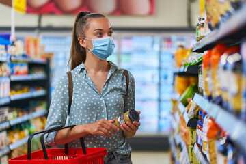 Young woman buyer wearing medical protective mask among the shopping shelves chooses food products...