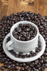 Top view of white coffee cup with roasted coffee beans on wooden table with more beans, selective focus, vertical
