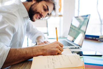 Meditative young man with stubble working at home in bright wooden room in his own house. Remote workplace. Smiling and writing with pencil