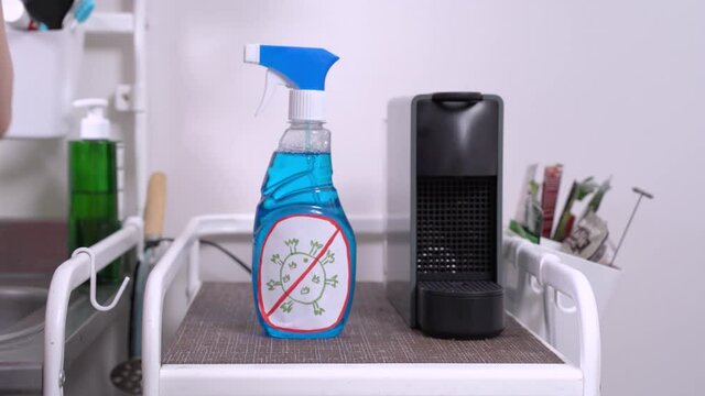 Human hand putting plastic bottle with blue transparent liquid inside, label with picture of virus molecule, on the shelf in the kitchen or housekeeping room. Cleaning home with antiseptic treatment.