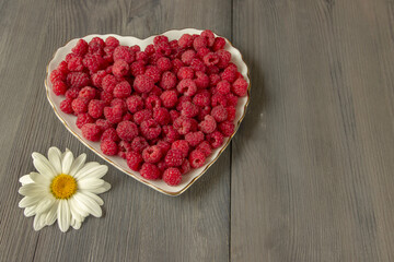 Chamomile flower and a dish of raspberries on a plate in the shape of a heart. The view from the top.