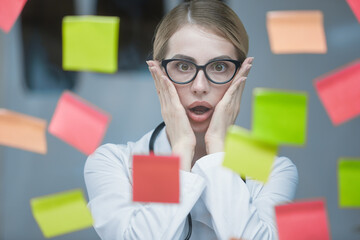 An emotional young doctor in the office, with a stethoscope around his neck and in a white coat, poses against a background of colorful sticky notes glued to a transparent glass.