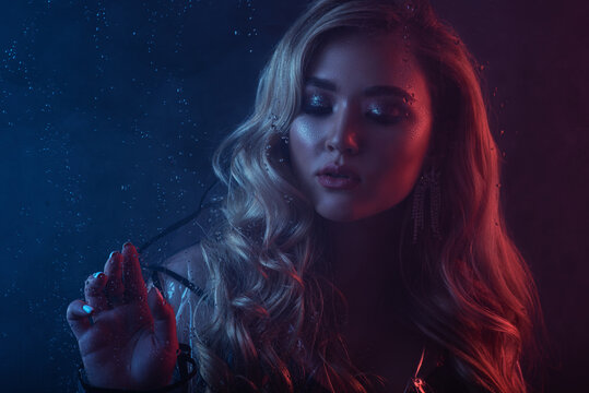 Fashion, beauty portrait of elegant blonde woman posing in colorful bright neon uv blue and red lights, smoke. View through the wet glass with rain drops. Copy, empty space for text