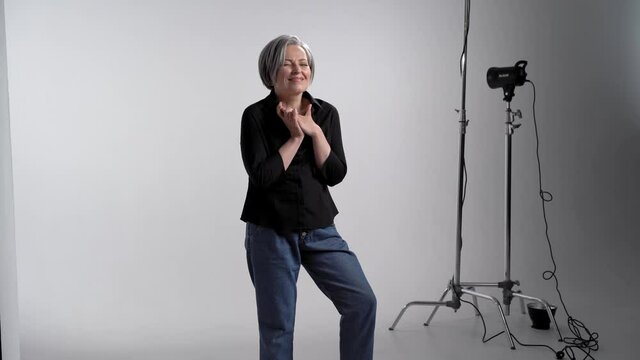 Dreaming adult gray haired woman showing facial expressions posing for photographer in studio on a white background in casual clothes black shirt and jeans with studio light equipment. Prores 422.