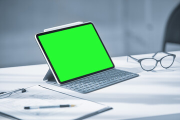 A tablet with a green screen on a white Desk in a white office, glasses and pen next to it