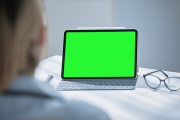 Medical worker a woman in a white coat uses a tablet in the office at the Desk, chromakey on the tablet screen, a view over her shoulder