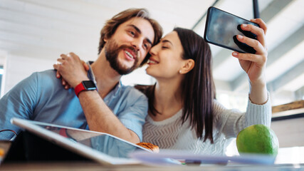 Couple working together at home in bright wooden room in their own house. Remote workplace. Taking photo using phone and kissing