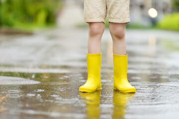 Little boy wearing yellow rubber boots walking on rainy summer day in small town. Child having fun.