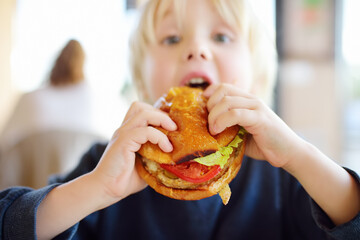 Cute blonde boy eating large hamburger at fastfood restaurant. Unhealthy meal for kids. Junk food.