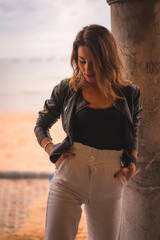 Lifestyle in the city with a blonde girl in white pants and a leather jacket near the beach. Perched by the sea looking at the ground