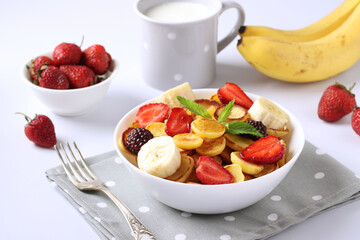 Trendy tiny pancakes for breakfast with strawberry, blackberry and banana in bowl on white background and cup milk. Closeup