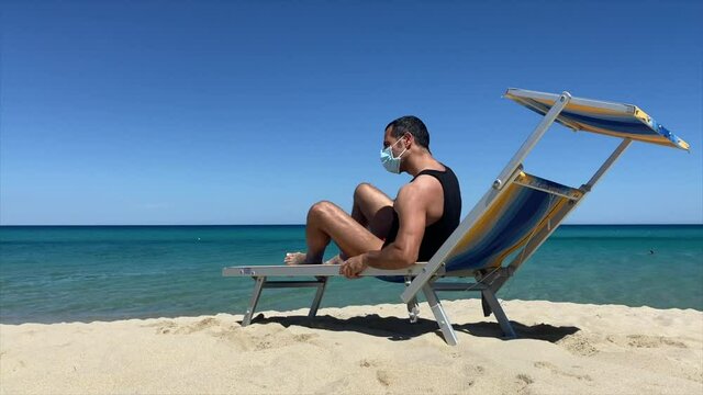Uomo moro prende il sole in relax in spiaggia al mare  con la mascherina facciale , sullo sfondo un cielo blu e un mare cristallino