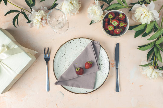 Flat lay composition with white peony flowers, empty plate with napkin and cutlery  on a light pink concrete background