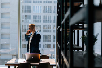 Fototapeta premium Professional young businessman wearing fashion suit is talking on mobile phone in modern office room near large window. Concept of office working.