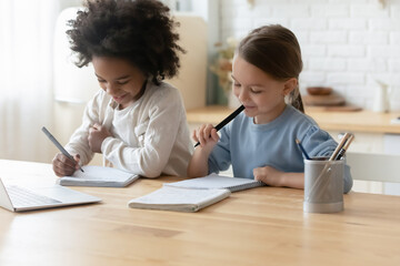 Two multiethnic sisters little girls doing homework sit in kitchen at home. Holding felt-tip pens writing essay on workbook, thinking on common task. Homeschooling, education, learning process concept - obrazy, fototapety, plakaty
